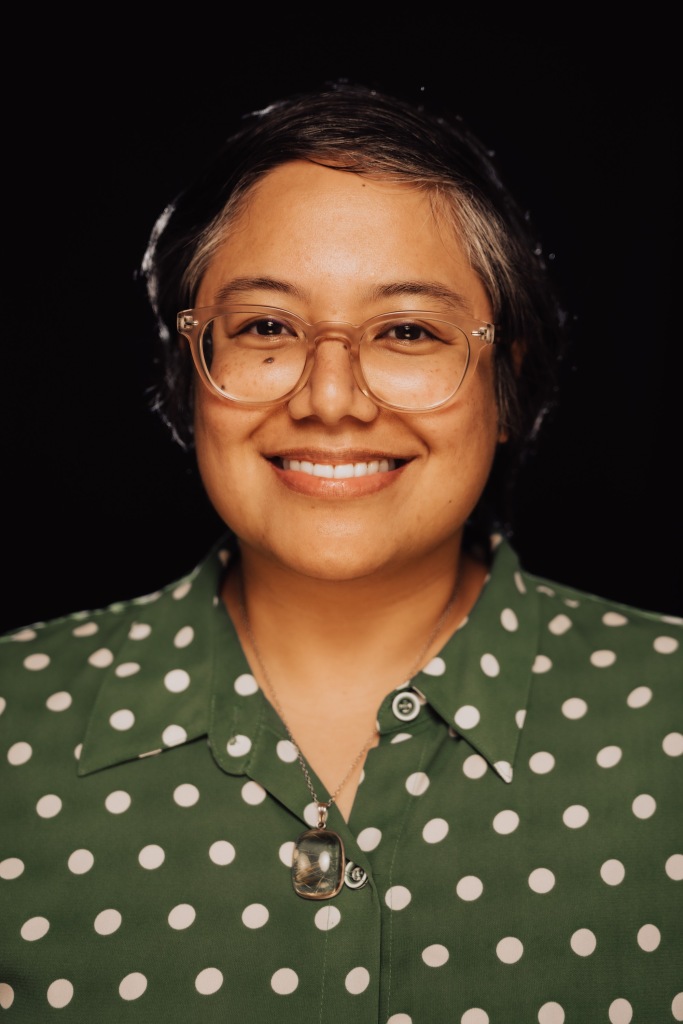 A smiling Filipino person with short hair wearing clear glasses, a green and white polka dot button up top, and a quartz pendant necklace.